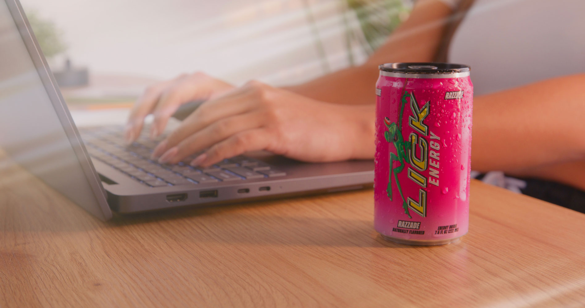 Person using a laptop with a pink Lick Energy drink can on a wooden table.