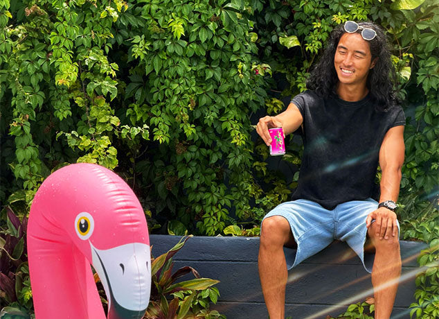A man sitting outdoors with a pink inflatable flamingo and a can of Lick Energy, surrounded by greenery.