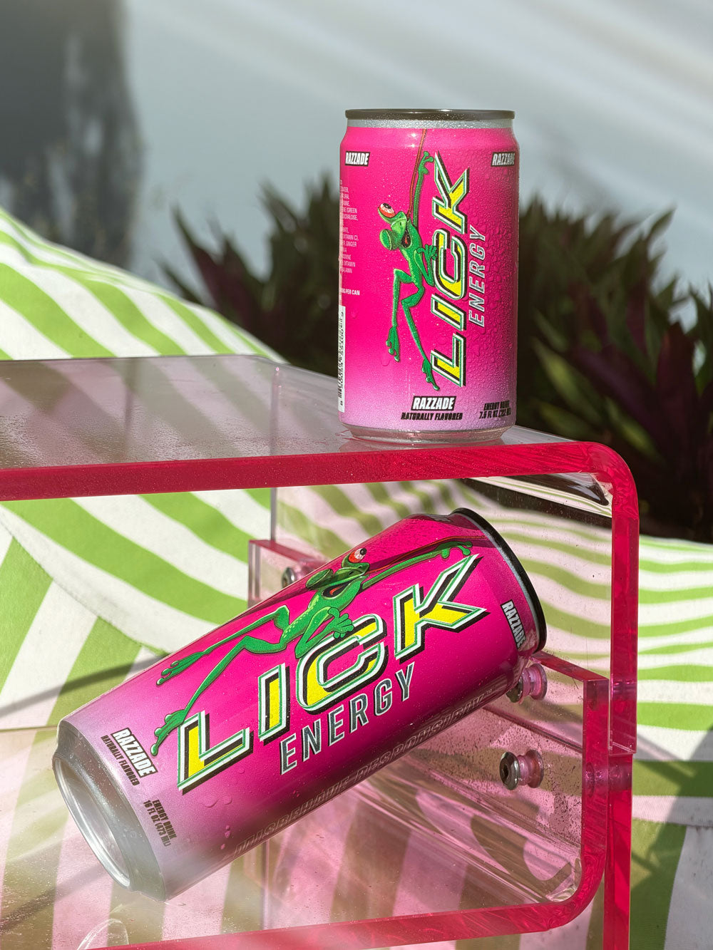Two pink Razzade Lick Energy cans on a transparent shelf with a blurred outdoor background.