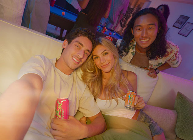 Three friends sitting on a couch, smiling and holding Lick Energy drinks in a casual setting.