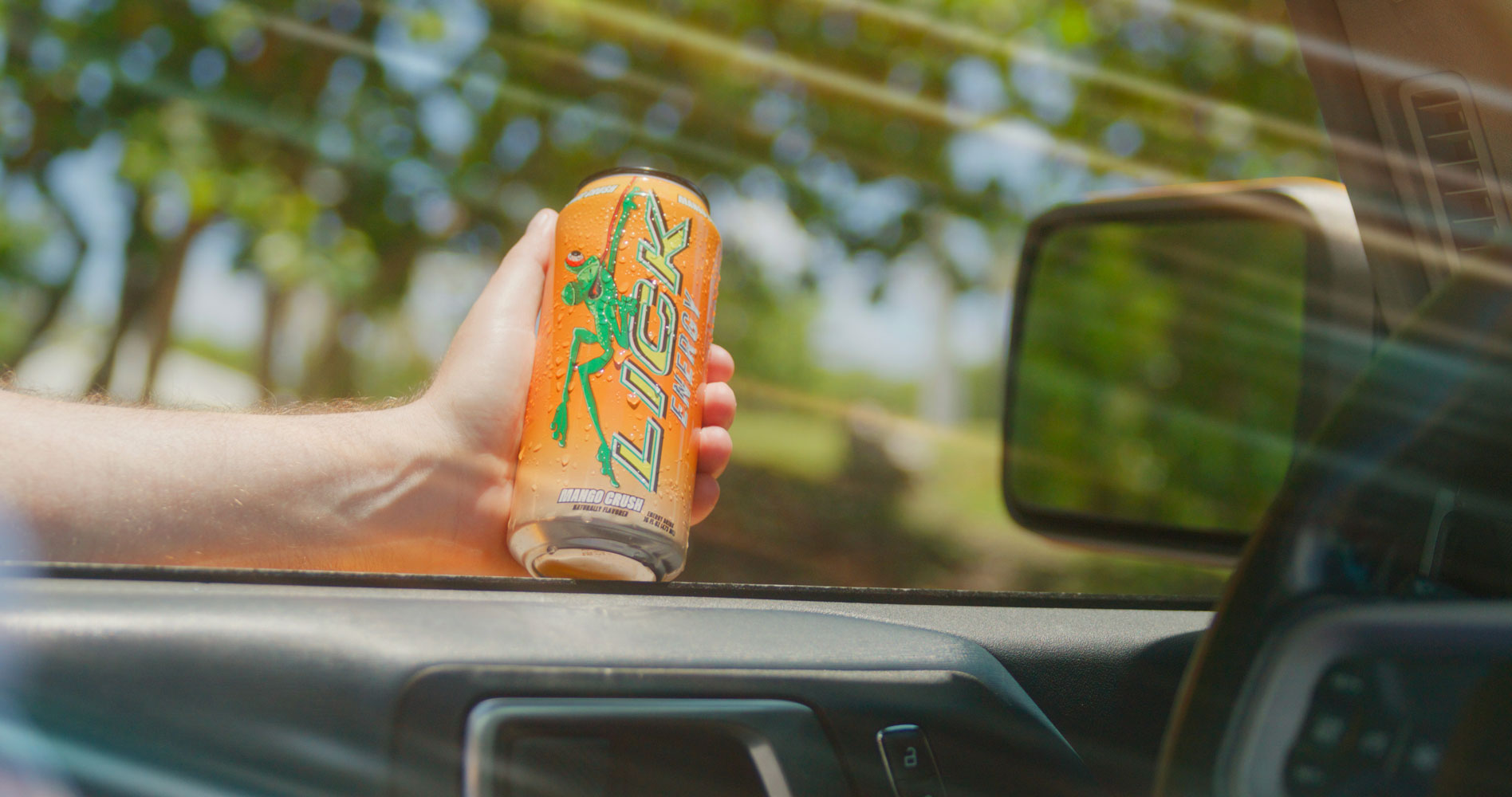 Hand holding a can of LICK Energy against a car's rearview mirror with a blurred outdoor background