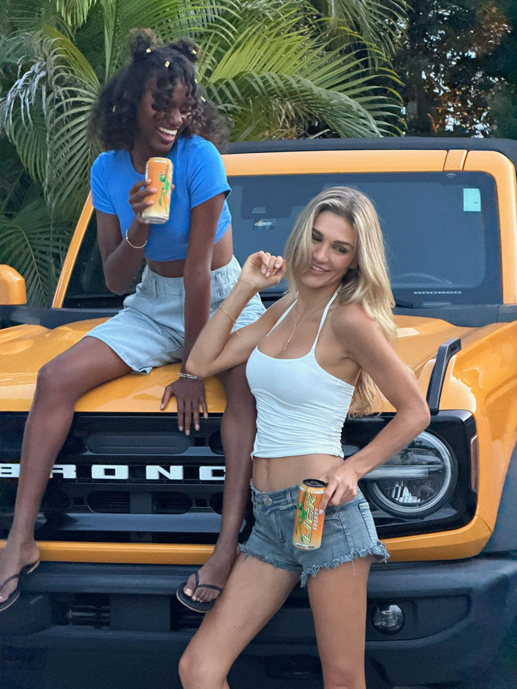 Two women sitting on a yellow Ford Bronco with drinks, palm trees in the background with LICK Energy Cans