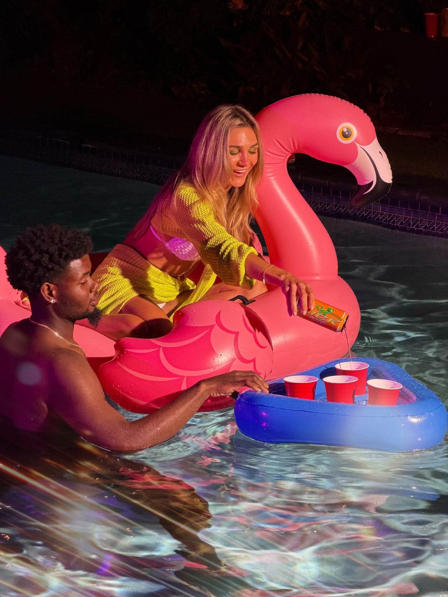 Two people on a pink flamingo pool float pouring Lick Energy into a red Solo cups in a pool.