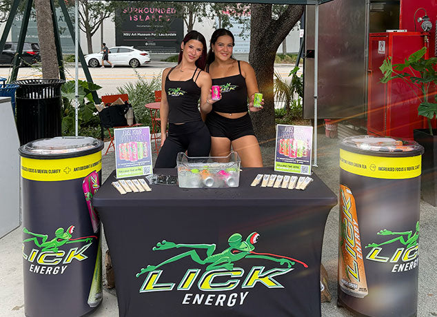 Two women standing behind a Lick Energy drink booth outdoors.