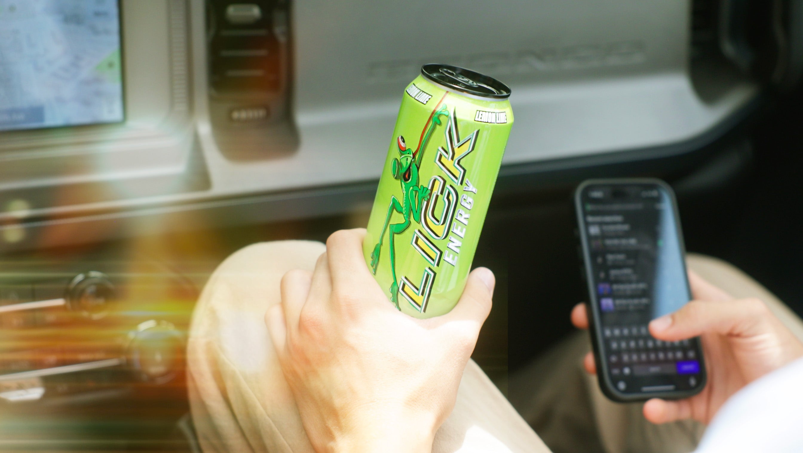 Person holding a can of LICK energy drink and a smartphone inside a vehicle.